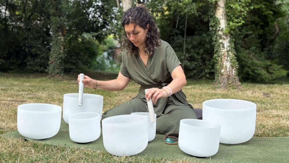 A woman with long brown curly hair, wears olive coloured robes. She is sitting cross legged on grass in front of several tress and is creating music from several white bowls in front of her of varying sizes.