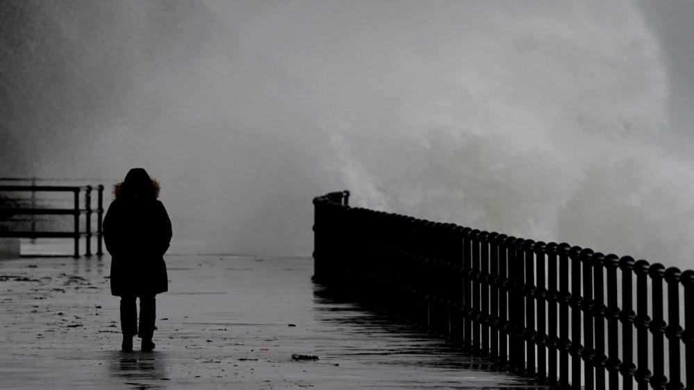 A silhouetted person walks along a paved seafront wearing a raincoat. To the right of the image, sea splash from a large wave spills over a silhouetted fence on to the pavement behind the person. Conditions appear wet, windy and grey.