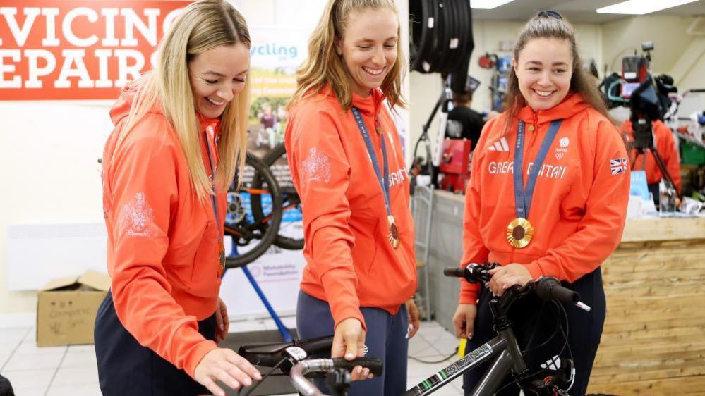 Olympic gold medallists Katy Marchant, Josie Knight and Sophie Capewell and orange jumpers smile and look at a bicycle