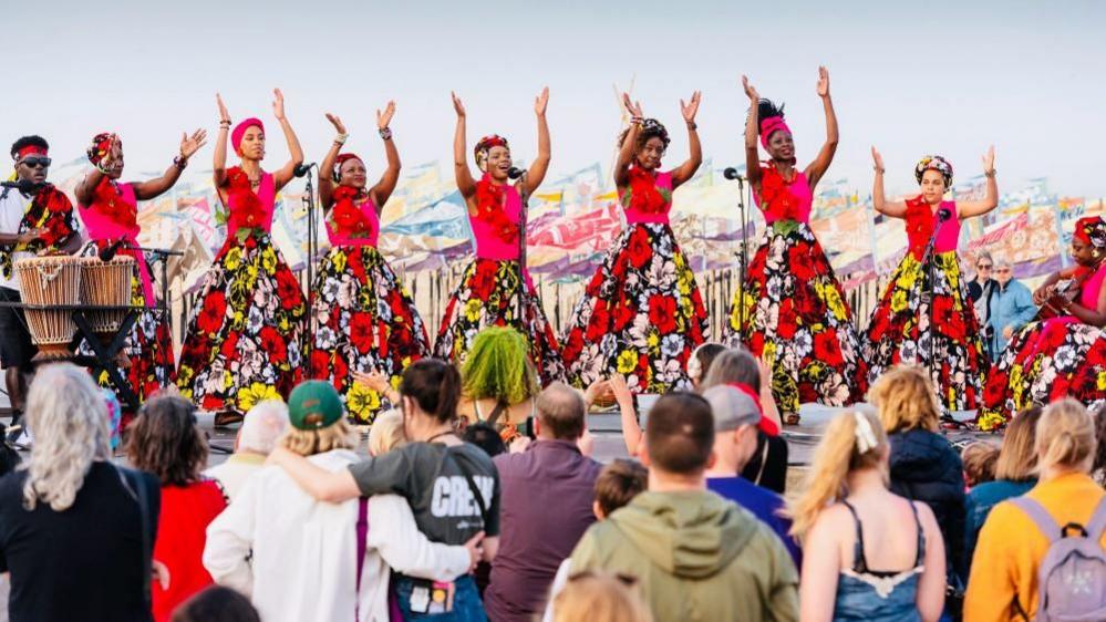 The African Choir of Norfolk, with its choir members dressed in magenta tops and hairbands, and colourful floral dresses. They are performing on the beach in Great Yarmouth with a vast collection of silk flags made by children fluttering behind them, and a crowd of people watching on. A drummer is to the left of the choir.