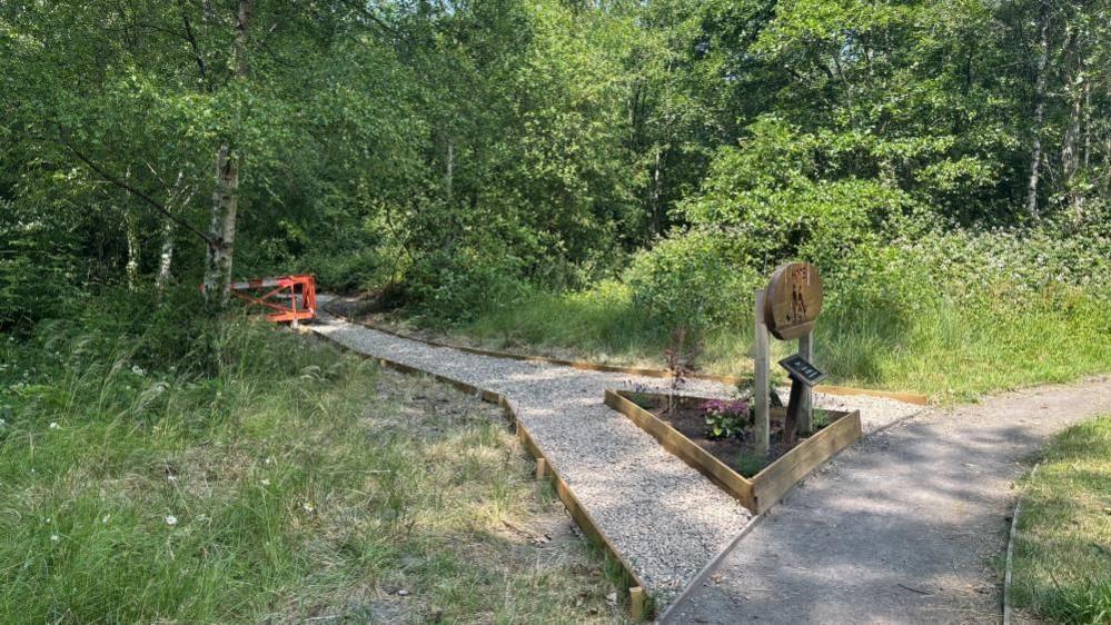 A stoney path that leads left through a wooded area. The area is full of trees and green shrubs. A sign and a flower bed are on the right-hand side of the image.