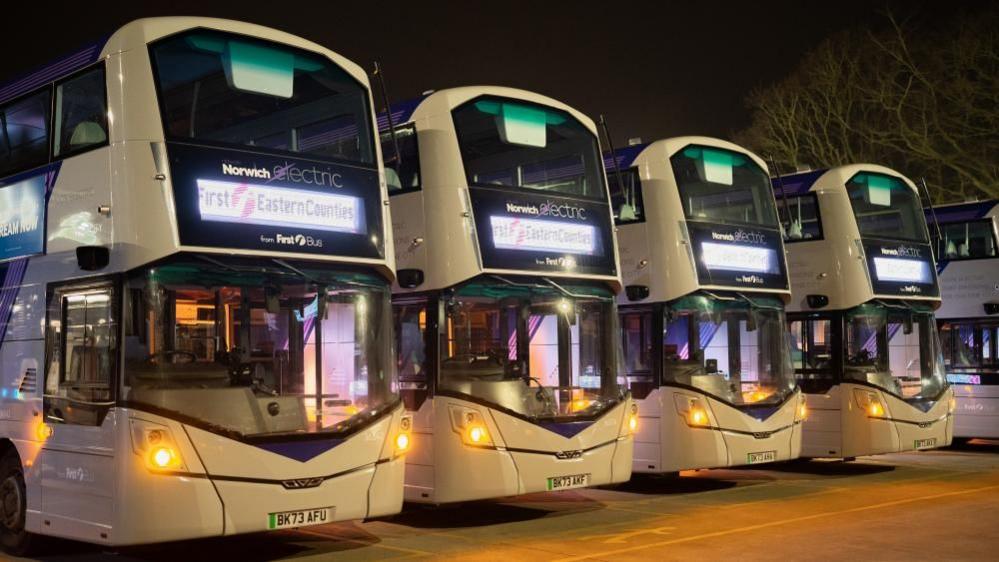 Five Norwich Electrics double decker buses are painted grey and purple, and feature an electric flash logo. The vehicles are standing in the depot in the night, with the area lit by street lamps. Each displaying First Eastern Counties with the First Group logo displayed on the destination board