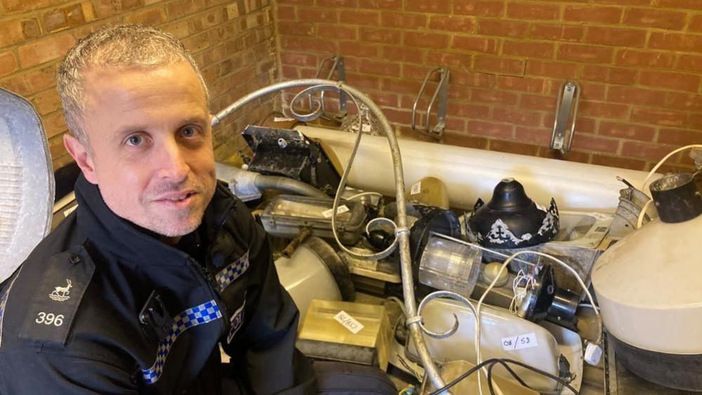 PC Tom Woollard is sitting next to a number of lamps and lanterns in a warehouse. He has short, fair hair and is wearing a police uniform