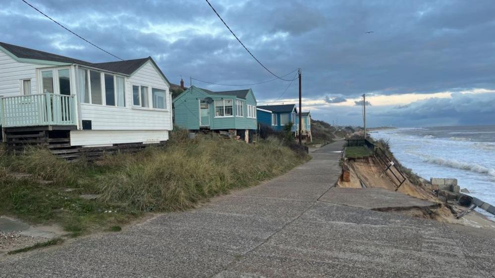 Wooden homes set back from concrete roadway which is being undermined by the sea.