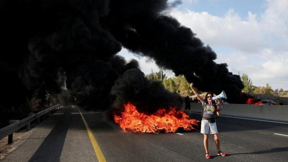 woman stands in front of fire on highway holding a megaphone