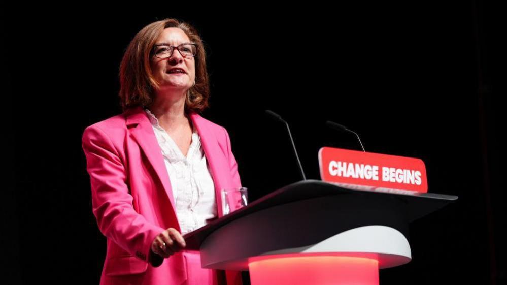 Eluned Morgan wears a pink suit and white blouse, with brown bobbed hair and glasses. She is speaking while standing at a lectern with the words 'Change begins' on it, at the 2024 Labour party conference.