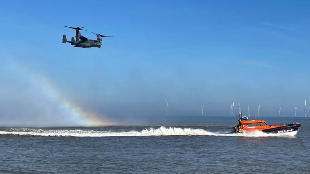 A USAF Osprey helicopter flies over the sea, with its downdraught causing a rainbow beneath it. Ahead of it is the Caister Lifeboat, with the wake trailing after the vessel