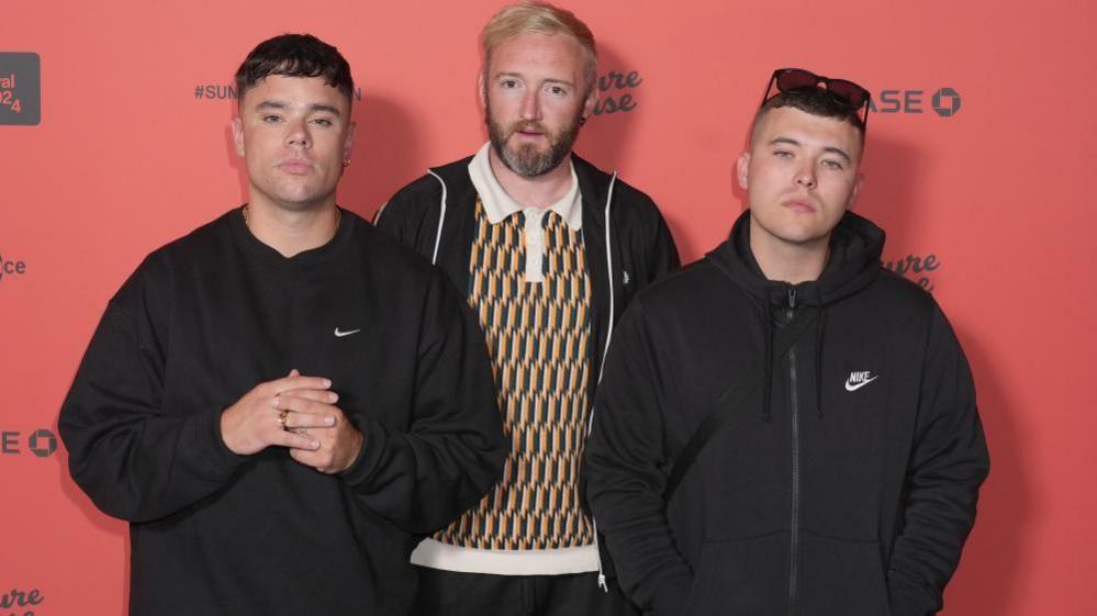 Mo Chara, left, is wearing a black sweatshirt and looking directly at the camera. He had short brown hair. DJ Provai, middle, is wearing a yellow and green patterned polo top and a black zipper jacket. He is looking directly at the camera and has a beard and ginger hair. Moglai Bap, is wearing a black zip up coat. He is wearing sunglasses on his head and has short brown hair.