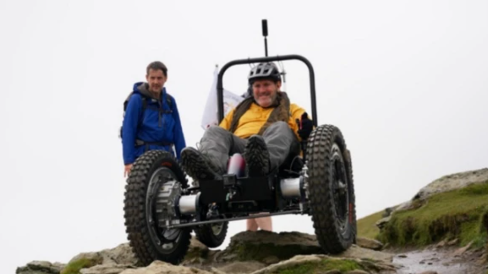 Nick Wilson in his RockClimber chair on a mountain top. It has very large tyres. He is wearing a yellow top and a helmet. Two people are watching, standing behind him.