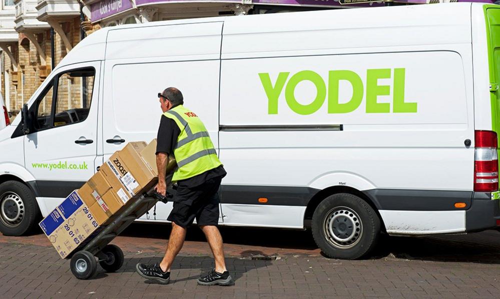 A Yodel parcel delivery driver walks in front of a white Yodel van with parcels in a reflective vest in 2018