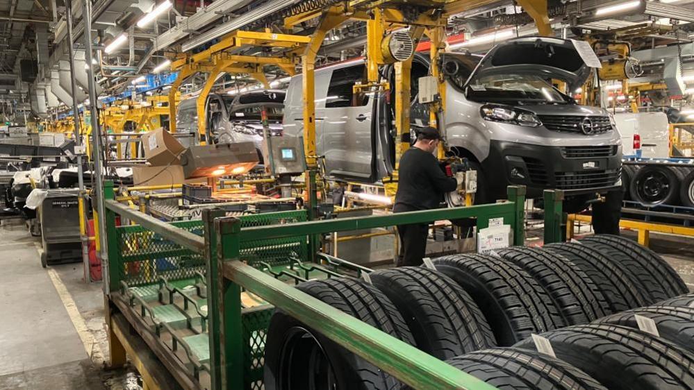 A worker in a Vauxhall Luton plant, working on a van, with tyres and car parts around him. There is a lot of machinery, hanging from the ceiling and more vans in a production line. 