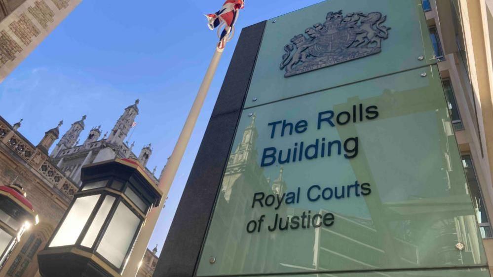 A white sign with the words "The Rolls Building Royal Courts of Justice" written on it in back. The to left of the sign is a brown building and a flag pole with a red, white and blue flag hanging loose at the top. Reflections of buildings can be seen in the sign.
