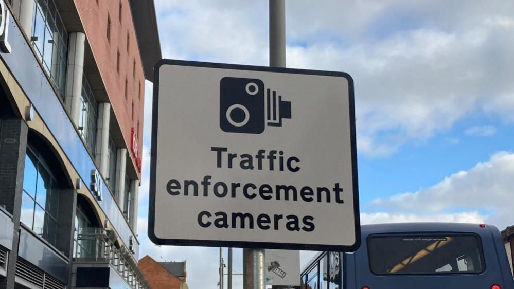 A "traffic enforcement cameras" sign. It features a picture of a traditional camera in black on a white background. A red and black building is to the left and a blue bus is to the right.