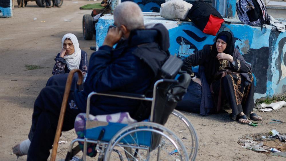 A man with an injured foot sits in a wheelchair parked on a dirt road. Two women sit either side of him on the ground