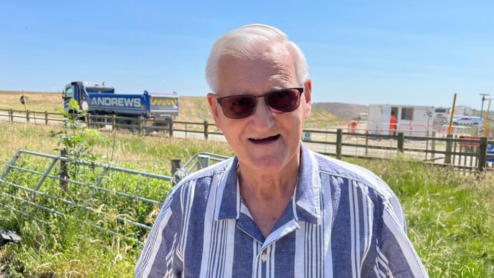 Peter Deeley with short white hair wearing brown sunglasses and looking at the camera in a blue and white shirt. He is standing in a grass field with a gate behind him. Beyond that is a wooden fence, behind which a blue lorry marked ANDREWS can be seen on a temporary road. There is a small white hut alongside the road.