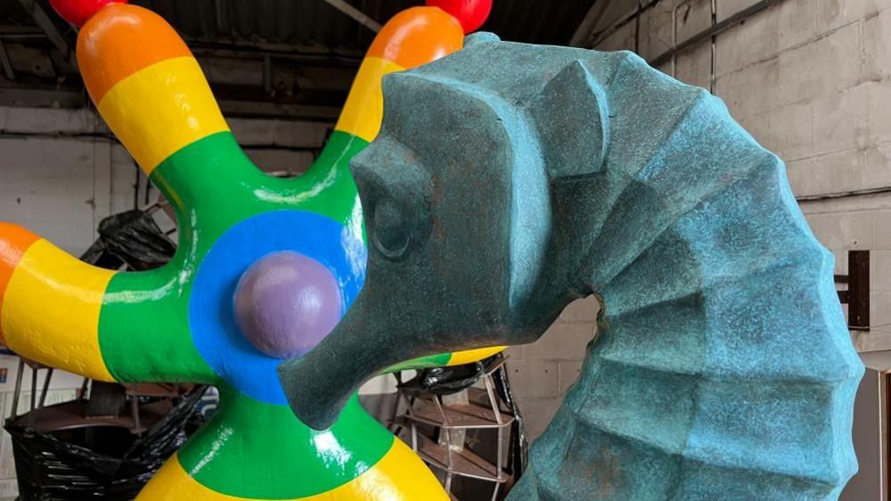 A sculpture of a seahorse, which is a mottled weathered copper colour, stands before another sculpture called The Juggler, which has two legs and four arms, painted in red, orange, yellow, green, blue and purple in concentric colour bands. 