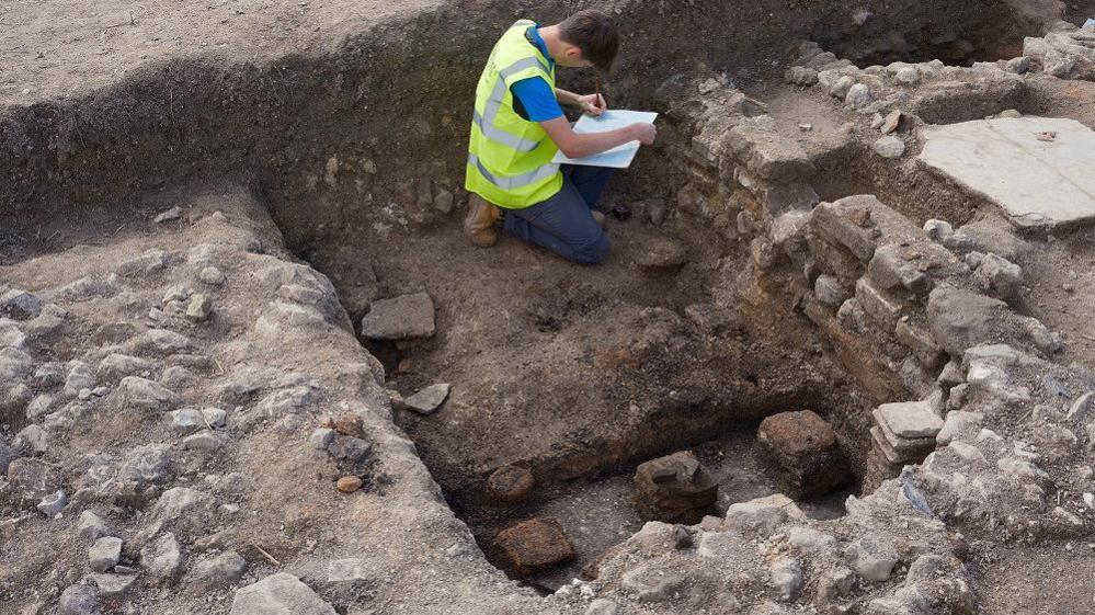 A volunteers records something on a clipboard while in a trench, which is quite deep and shows some stacks of tiles evenly spaced out, which would have been the hypocaust heating system.