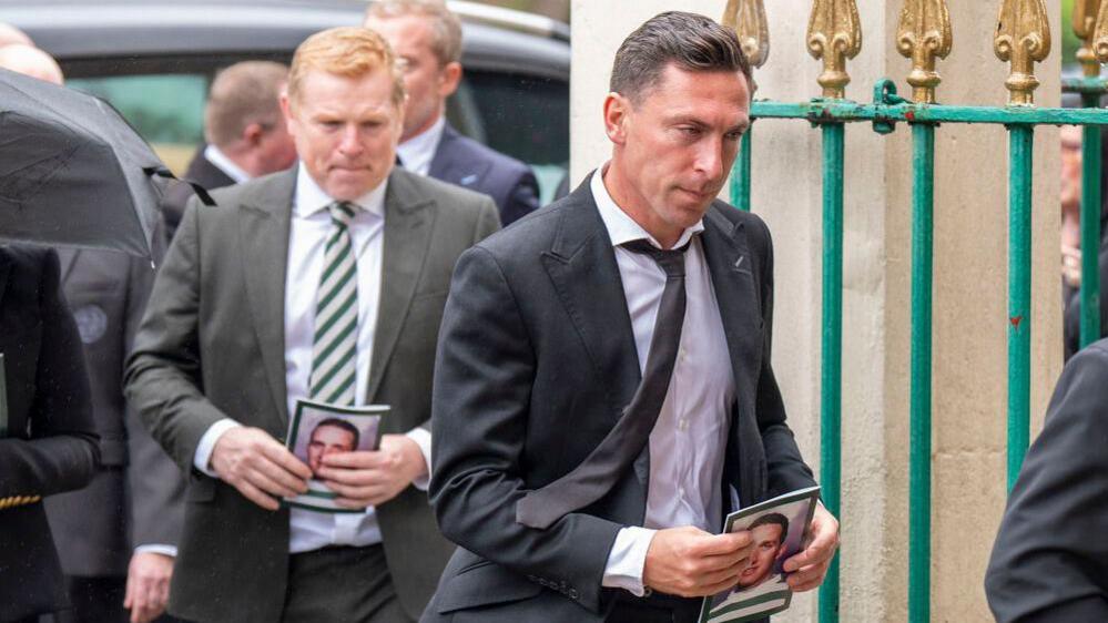 Neil Lennon and Scott Brown walk one after another wearing dark suits and holding funeral programmes with John Clark's face on the front