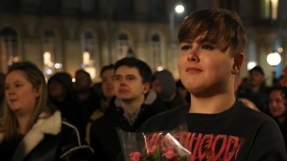 A young man in a black t-shirt holding a bunch of roses stands in a crowd with tears in his eyes during a vigil.