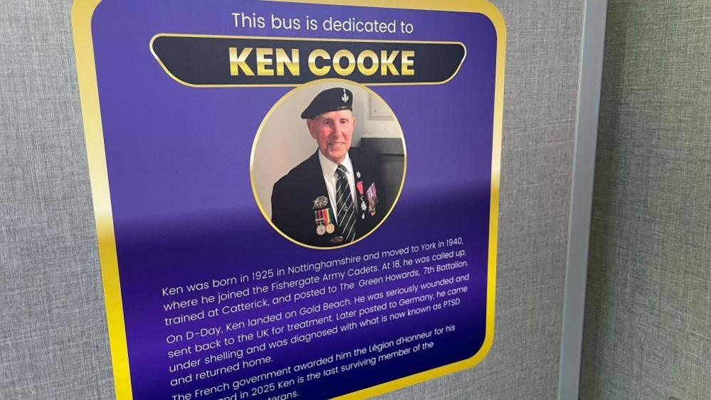 A purple plaque on a grey wall reads 'This bus is dedicated to Ken Cooke'. It has details about Ken's life written underneath and an image of an old man wearing a military uniform and medals. 