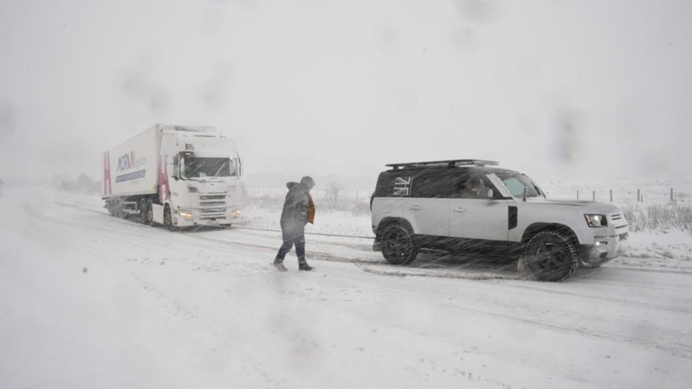 A 4x4 vehicle tows a lorry in snowy conditions