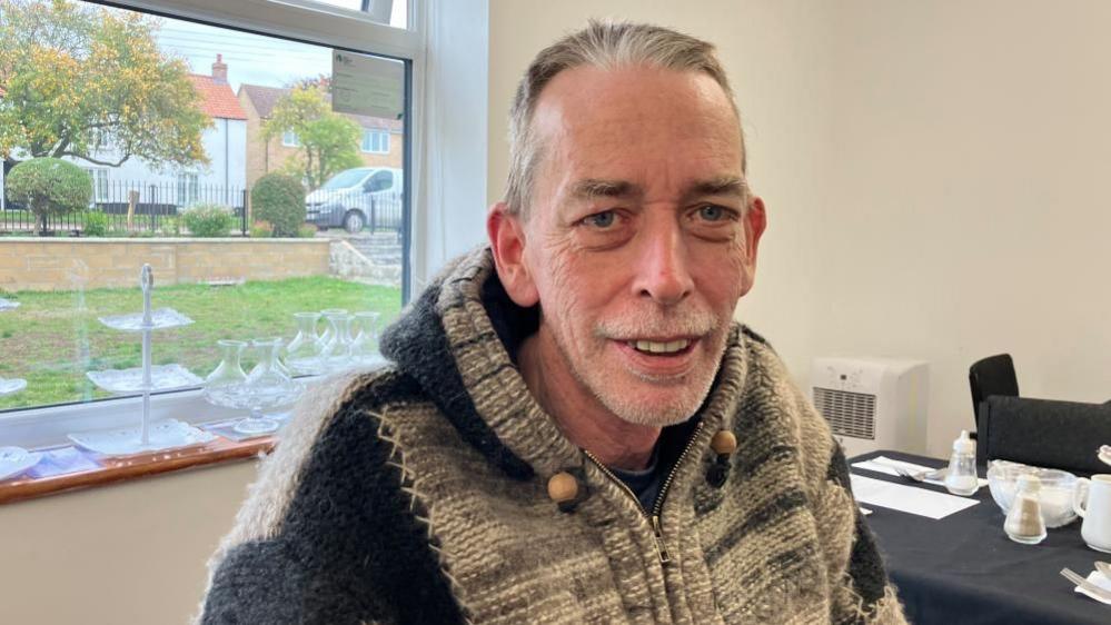 David Beek is sat next to window at a table with a view of a front garden. He is wearing a thick, hooded poncho-style jumper in shades of grey and beige. On the table beside him are several white ceramic cups, saucers, and a salt and pepper shaker. 