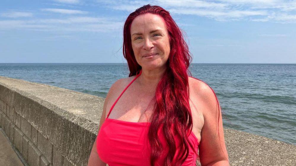 A woman in a neon-pink swimsuit smiles at the camera, with a view of the sea behind her. She has long, dyed red wavy hair.