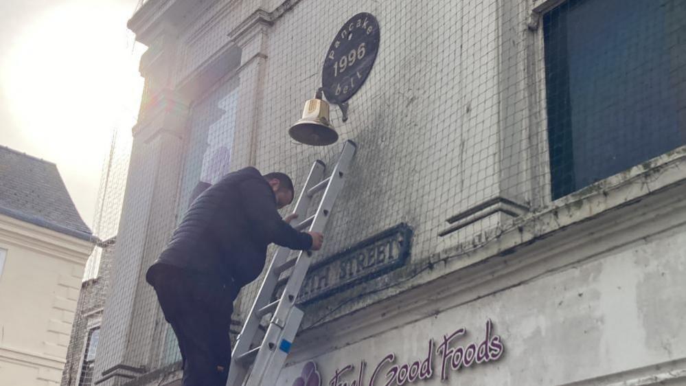 A man in a black jacket stands on a ladder which is leaning against a wall. The wall has a large gold bell attacked to it, with a sign that reads "pancake bell, 1996" above it. 