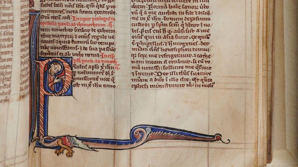 An old manuscript page, in tiny, perfectly written medieval Latin. One starting letter to a Bible book - a P - is lavishly decorated in blue and red and shows man inside the letter holding a book while the letter tails off dramatically, a mythical creature incorprated.