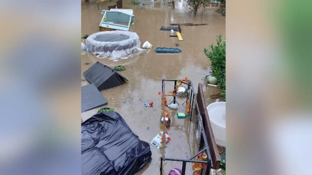 A view from above of a badly flooded garden. Trees can be seen submerged in deep brown water. A shed is floating on its side in the water, along with other garden furniture and debris.