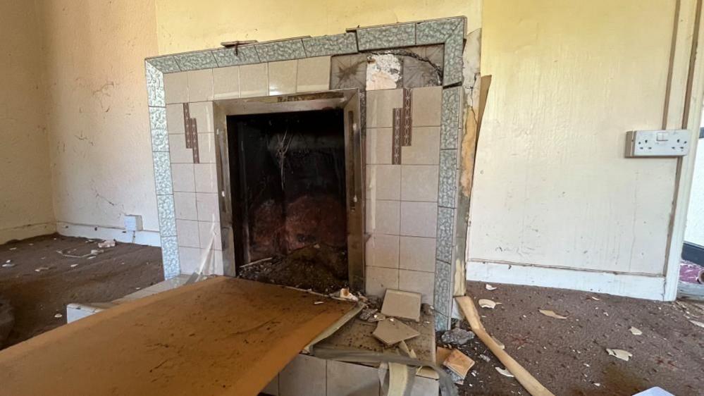 A tiled fire place, damaged with several tiles missing or broken. A board that once covered the fire aperture, lays horizontally. There is detritus and dirt on the carpet and the wall appears dirty. A double socket is mounted on the wall nearby.