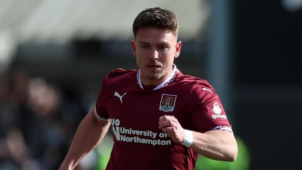 Sam Hoskins wearing a claret Northampton Town kit running.