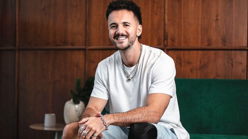 Luke Tarrant, sitting on a green sofa, looking at the camera and smiling. He is wearing a white top, with several silver necklaces, rings and a silver bracelet. He has shorts on, you can see one knee and the start of an artificial leg.  