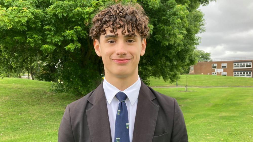 Aidan a pupil at Wootton Upper School wearing a dark school uniform, including a tie and blazer, standing in front of his school, with a tree and the school behind him