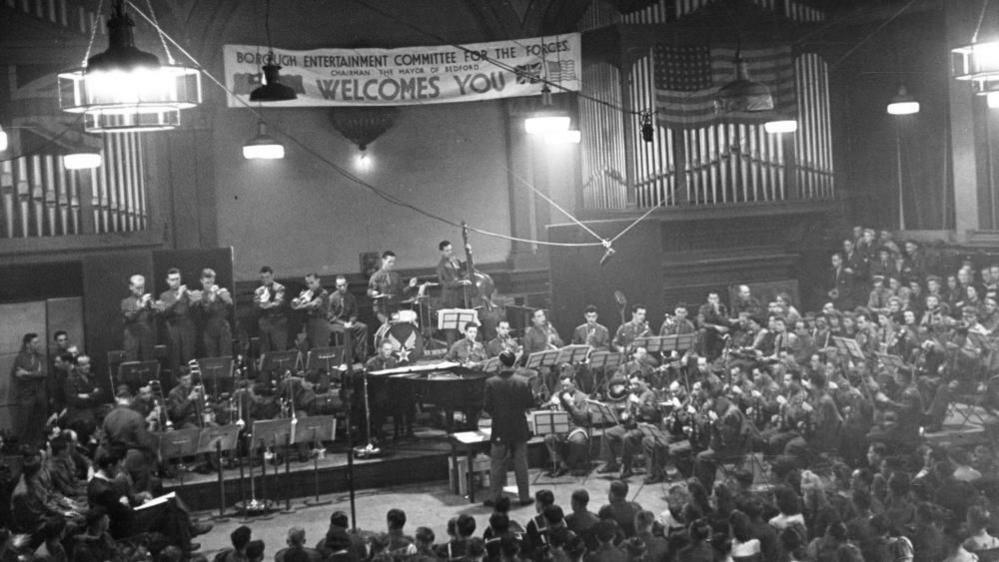 A black and white image showing Glenn Miller has his band in Beford's Corn Exchange, a banner is on the wall, there are organ popes and a very large orchestra, and people sitting down to watch them. 