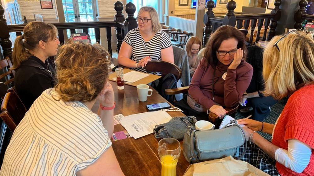 Group of women sitting around a pub table.  They are talking to each other and some are taking notes. Some have mugs while others have soft drinks in glasses.