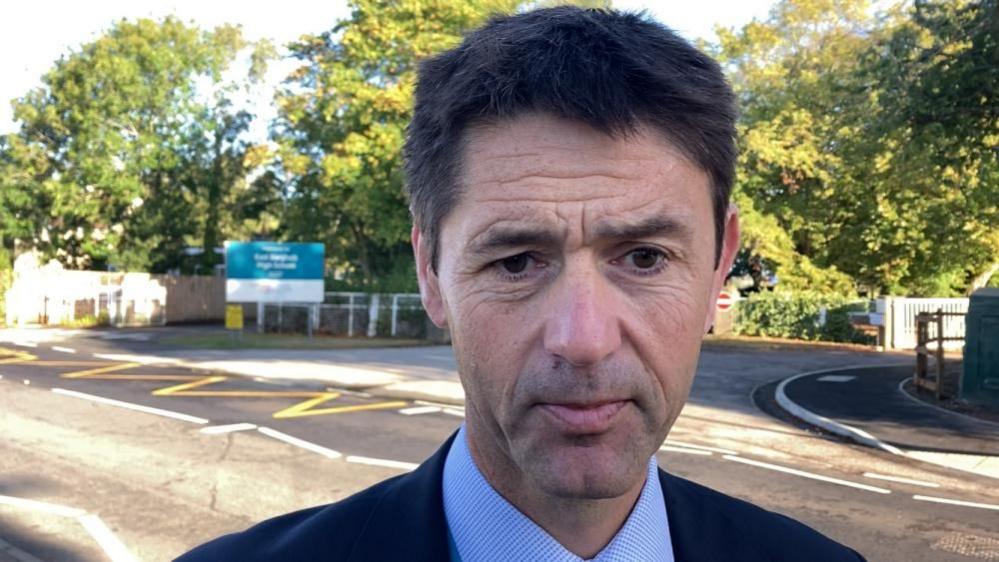 Dan Woodcock, a head teacher, standing outside his school, wearing a suit and shirt, looking concerned and to the right of the camera 