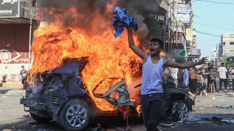 Palestinian boys reacts in front of Israeli car on fire in Gaza (07/10/23)