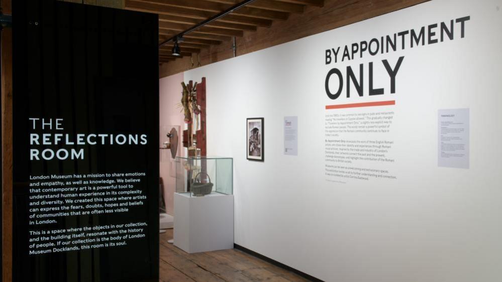 The entrance to the Reflections Room with By Appointment Only printed on the white back wall. Art pieces can be seen in the background in glass cabinets.