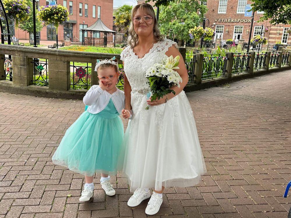 Claire and Emilia pictured on Claire's wedding day. Claire is wearing a white, tea-length wedding dress with a lace v neck top half and a diamante belt, with an a-line tulle skirt. She has white trainers and is holding white flowers. Emilia is in a turquoise bridesmaid dress with a tulle skirt, white bolero cardigan and white trainers. They are pictured outside on a paved area with gardens behind them.