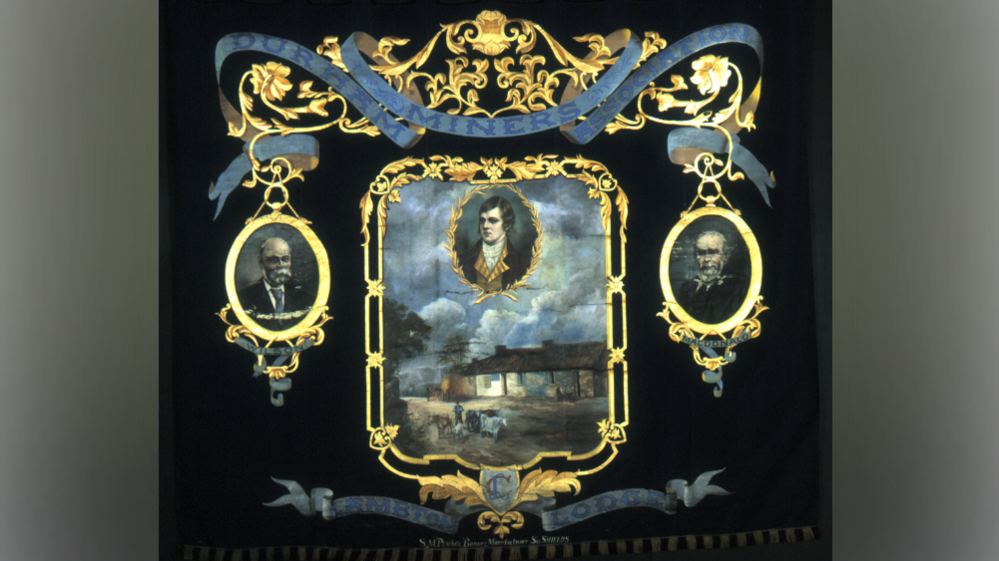 A black , blue and gold banner. It has a gold framed portrait shape in the middle with a painted portrait of Robert Burns and an image of mining lodges and a man with farm stock underneath. Two other portraits of J Wilson and A Macdonald are in a gold circular emblems to the right and left. On a blue ribbon it reads 'Durham Miners Association' 'Lambton Lodge'