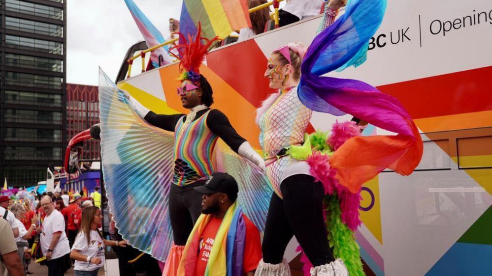 Two women on stilts standing in front of a bus, which they are nearly as tall as. They are dressed in costumes that have multi-coloured wings and brightly-coloured unitards with black leggings underneath. They have one arm in the air as well as Pride flags painted on their face.