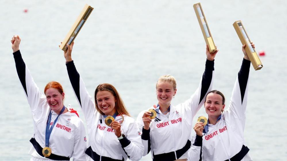 Four women in white GB team outfits hold their medals and medal tubes aloft.