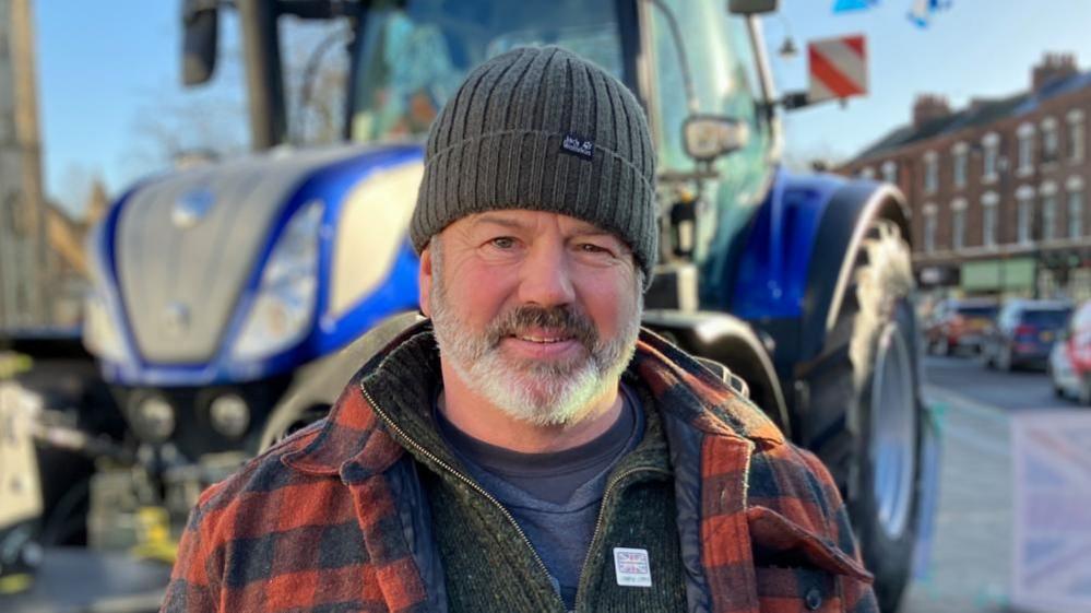 Richard Bramley is smiling looking straight ahead. He is wearing a dark brown hat with a red and black checked shirt with a jumper underneath. In the background there is a blue tractor.