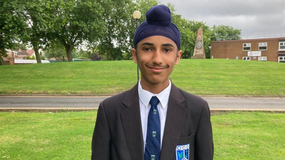 Manroop smiles at the camera as he is photographed outside the school building, he is wearing school uniform of a blazer, shirt and tie. 