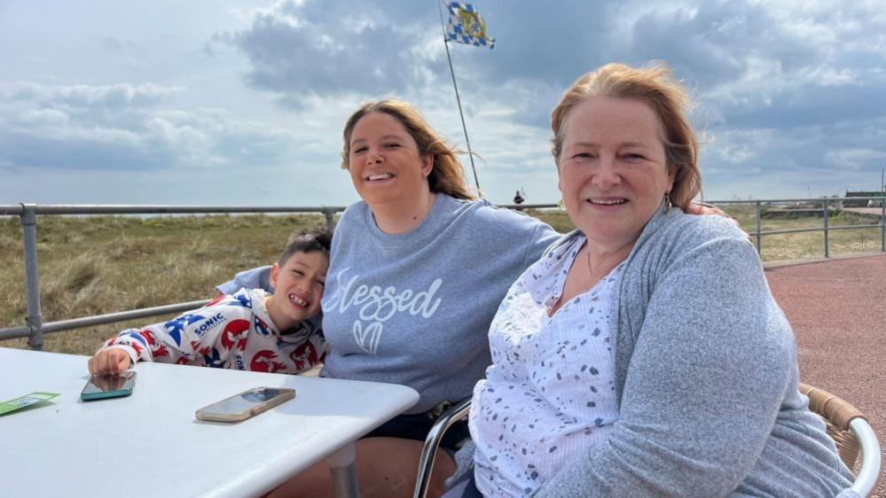 Cindy Gough, on the right, wearing a white spotty-patterned top, with grey knitted cardigan, has long strawberry blonde hair. She is sitting next to her daughter, Emily, who is wearing a grey sweatshirt, with printed words on the front. Her hair is auburn and below the shoulder. Her son, Cindy's grandson, Anakin, is on the left of the image, cuddling into his mother. He is wearing a red and blue patterned Sonic the Hedgehog top. They are sat at a table at Munchies Cafe on Great Yarmouth's north beach.