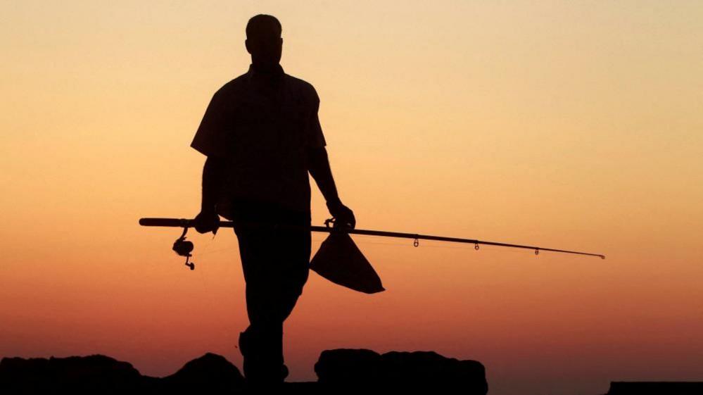 A man is seen in silhouette walking past some rocks. He appears to be wearing a short-sleeved shirt and trousers. He is carrying a fishing rod and a small bag. The sky is orange and yellow behind him