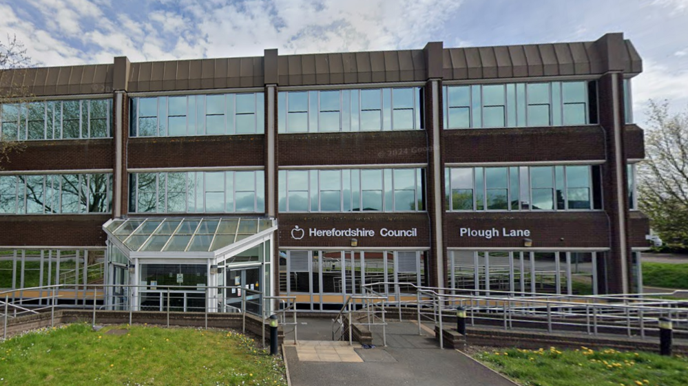 The front of Herefordshire Council's building. It is brown with several large glass windows. There are metal handrails outside the front doors and green grass