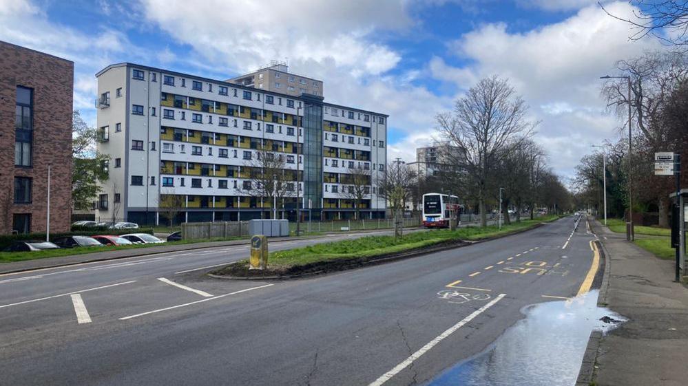 A road in Edinburgh bordered by flats. A bus stop can be seen on one side. It is a bright but cloudy day. 