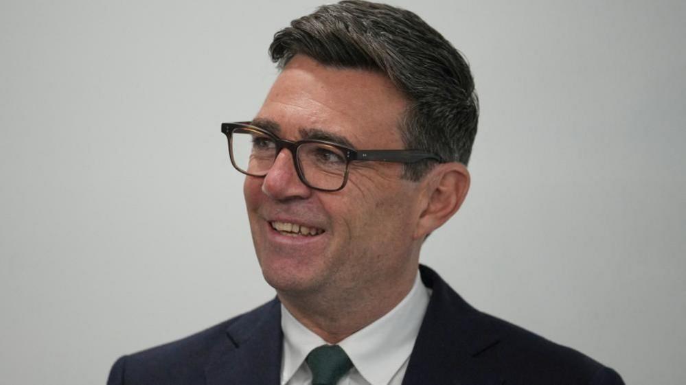 Andy Burnham looks off camera to the left. He is wearing a dark suit, white shirt and dark tie. 